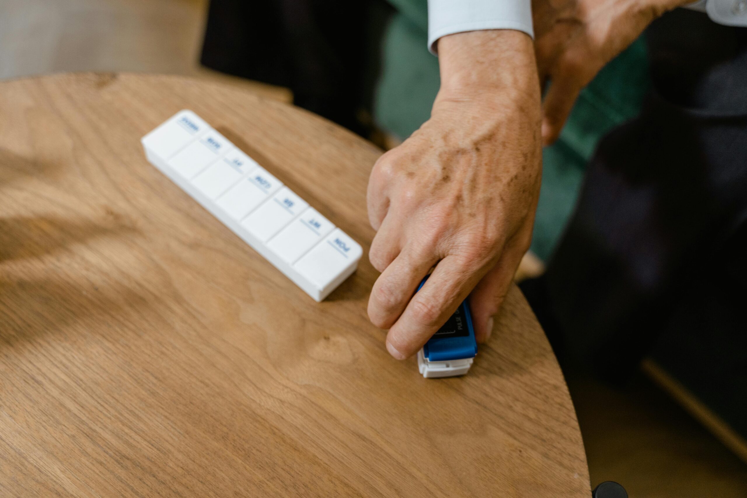 Close-up of an elderly person's hand using a pulse oximeter beside a pill organizer on a table.